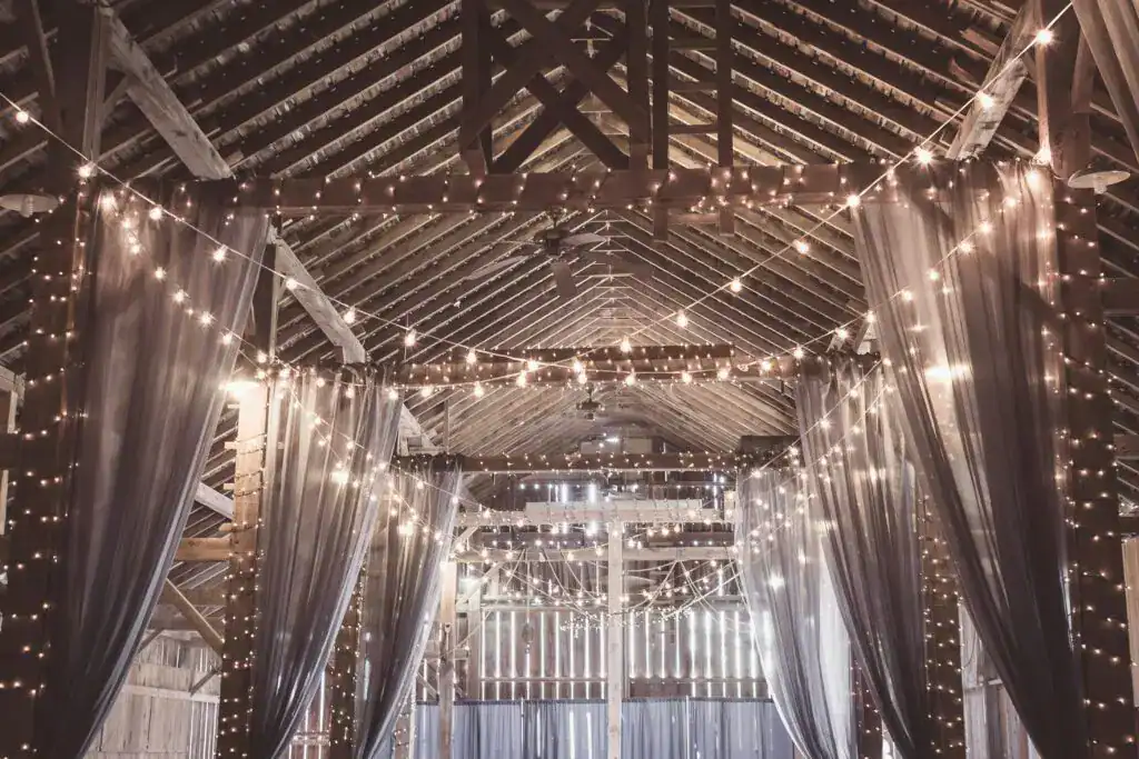 A rustic barn decorated with string lights and sheer drapes. The warm glow creates an inviting ambiance, highlighting the wooden beams and open space.