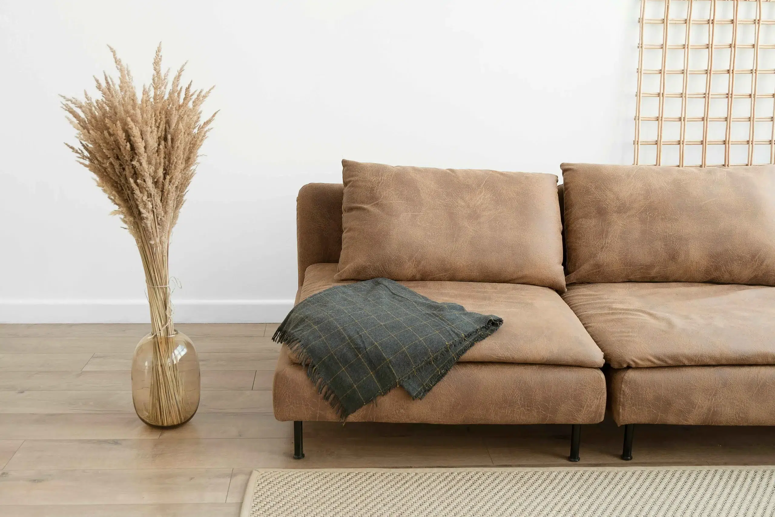 A cozy brown sofa with a dark plaid blanket draped over it sits next to a tall vase of dried pampas grass on a wooden floor, creating a warm, rustic atmosphere.