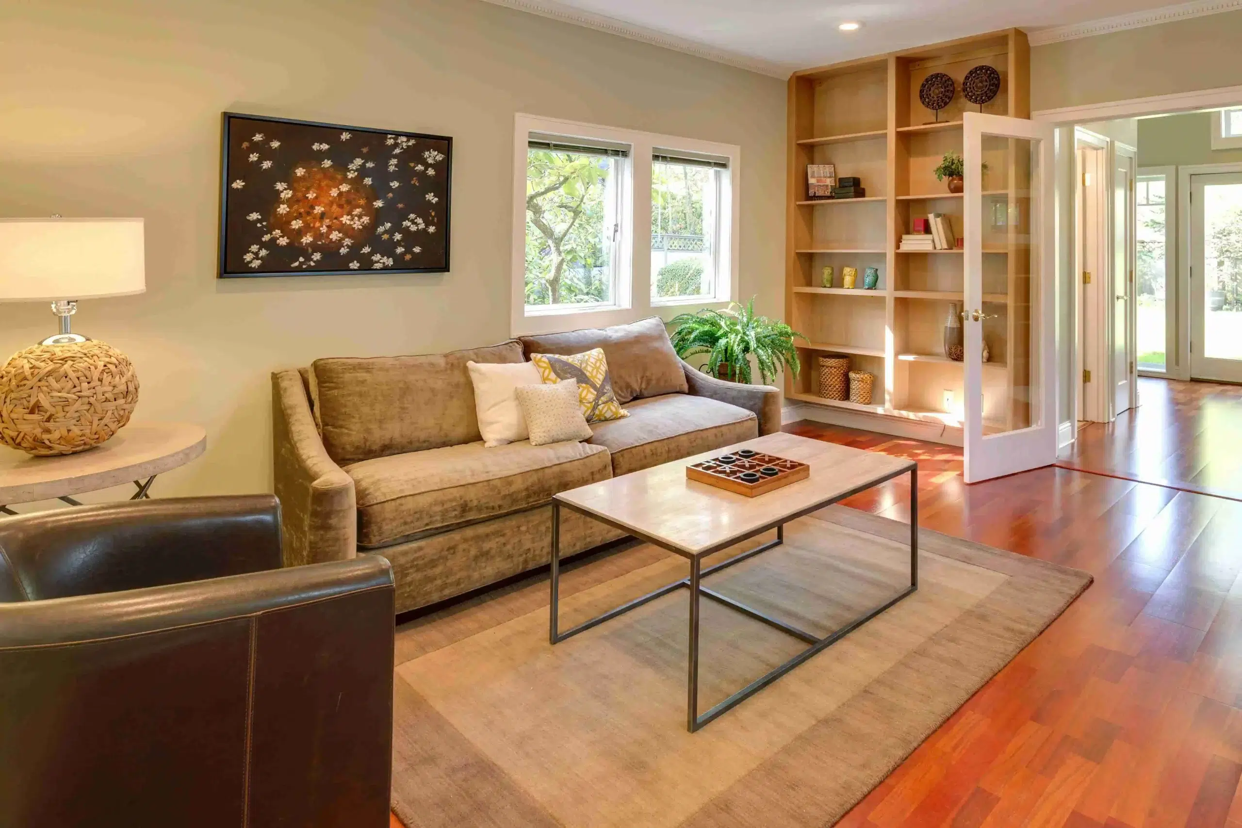 Cozy living room with beige sofa, patterned pillows, and a sleek coffee table on a rug. Wooden shelves hold decor, with natural light from large windows.