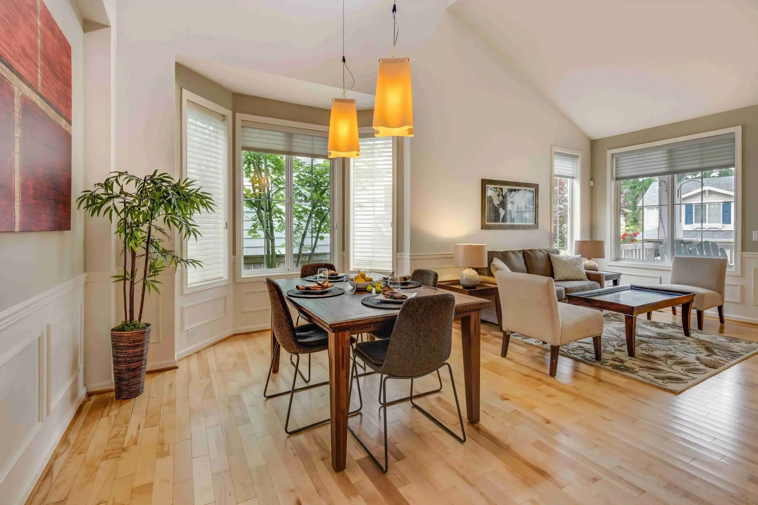 Bright open-concept dining and living area with wooden floors, a dining table set for four, modern pendant lights, a cozy sofa, and large windows.
